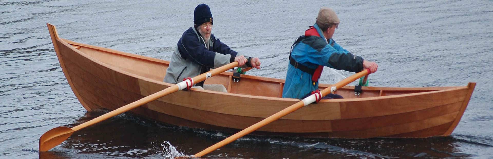 Two men in wooden rowboat on a lake, rowing.