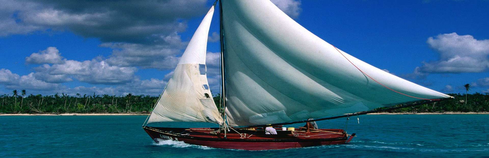 Beautiful wooden sailboat with sails catching the wind on shallow ocean waters by island shore.
