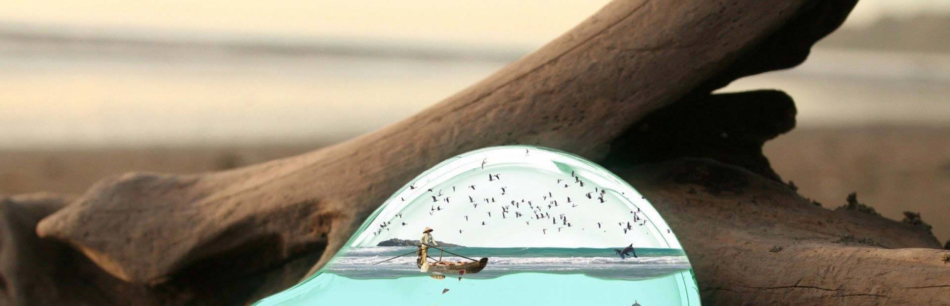 Piece of driftwood in background with surreal glass bottle in foreground with a man rowing a boat on an ocean contained within the bottle.