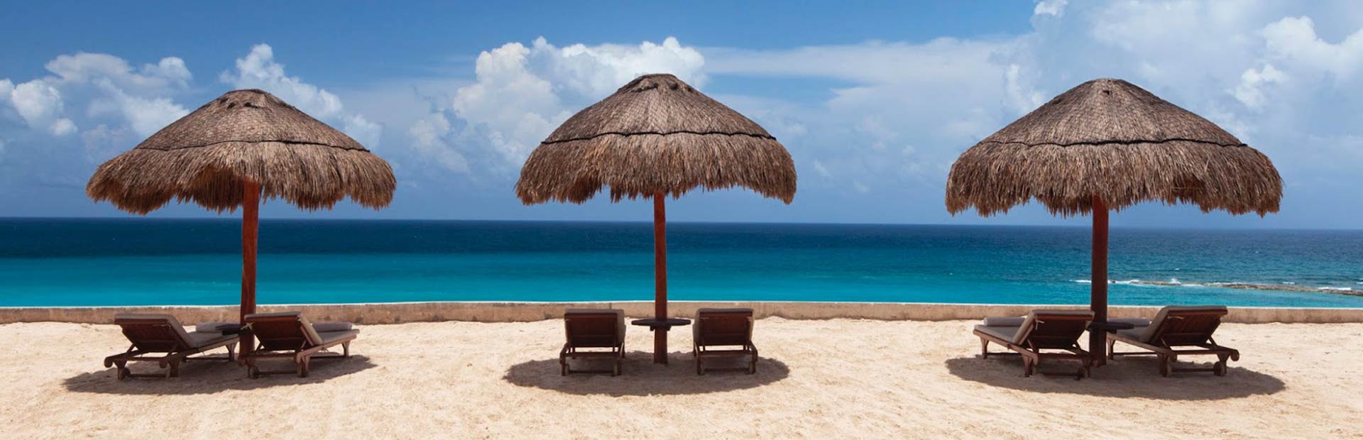 Three grass thatched umbrella shading areas with lounging seats on sand in front of the ocean.