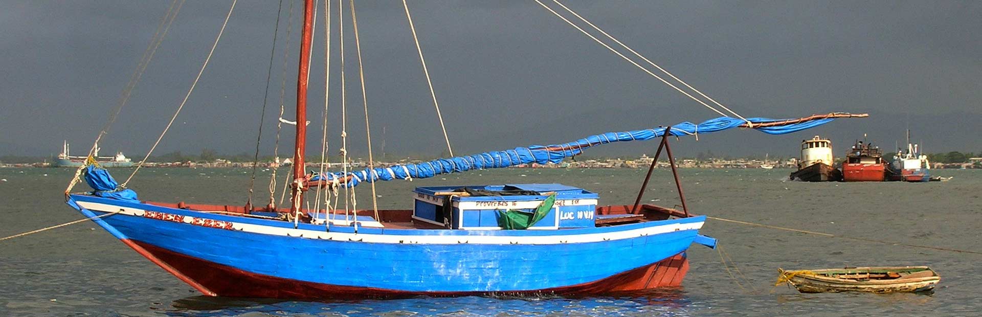 Large blue sailboat with sails down in foreground and fishing boats in the background offshore.